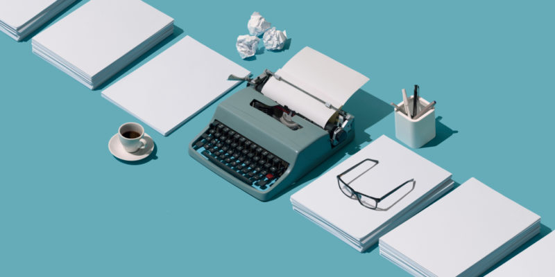 typewriter surrounded by paper, a coffee, glasses, and pens in a jar