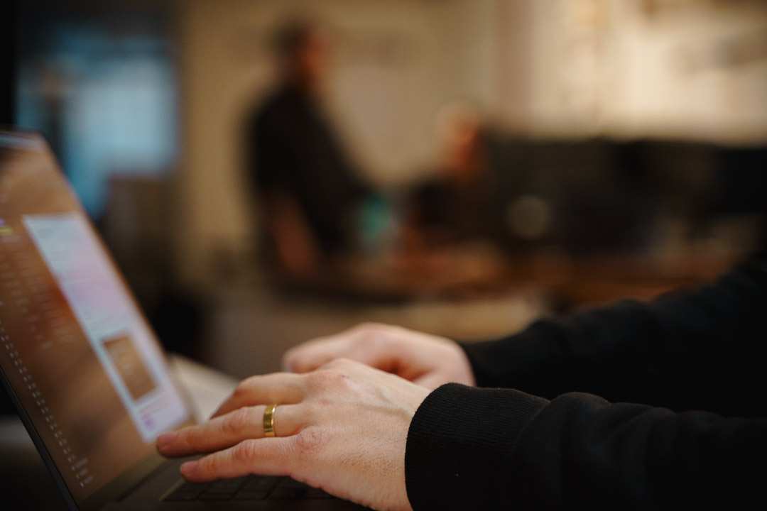 Close-up of hands typing on a laptop keyboard, with a gold ring visible on one hand. The screen displays a blurred interface with pink and white elements. The background is out of focus, showing two people standing and conversing in an office environment.